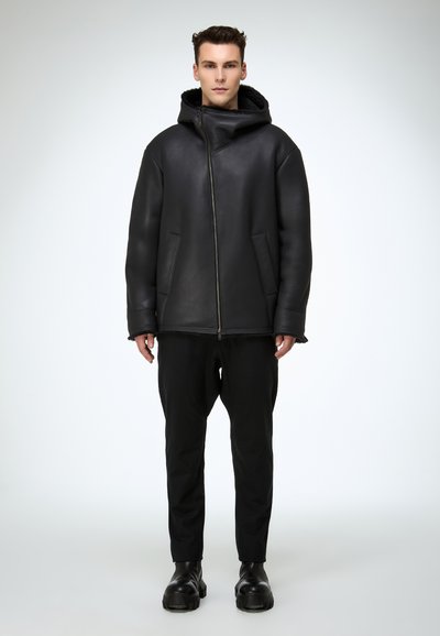 Schwarze Lederjacke mit Kapuze, Reißverschluss vorne und Seitentaschen. Getragen über schwarzen Hosen und klobigen schwarzen Stiefeln. Glatte Textur, lockere Passform.