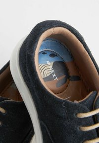 Svarta mockaskor med rund tå, beige snörning och en strukturera tanfärgad innersula med ett blått grafiskt mönster. Den vita sulan ger kontrast.