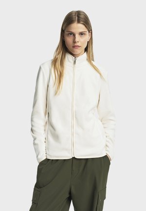 Veste en polaire blanche avec un col haut, fermeture éclair à l'avant et poches latérales. Associée à un pantalon cargo vert, montrant une coupe décontractée.