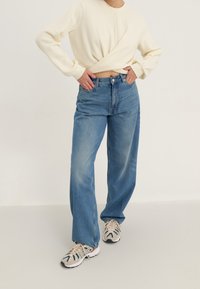 Felpa color crema chiaro, jeans a gamba larga a vita alta, blu slavato, tessuto strutturato, sneakers chunky con motivo geometrico in tonalità neutre.