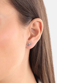 Orecchino a forma di infinito in argento con piccolo accento di gemma trasparente, posizionato su una tonalità di pelle chiara in vista profilo, evidenziando il lobo dell'orecchio.