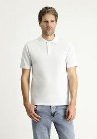 Polo shirt bianco realizzato in un tessuto testurizzato e traspirante, con una chiusura a tre bottoni e maniche corte, abbinato a jeans azzurri chiari.