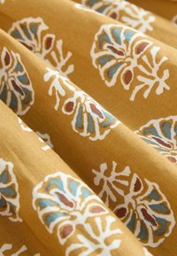 Tissu à fond jaune moutarde avec des motifs floraux blancs agrémentés d'accents bleus et bruns. Texture lisse, partiellement drapée.