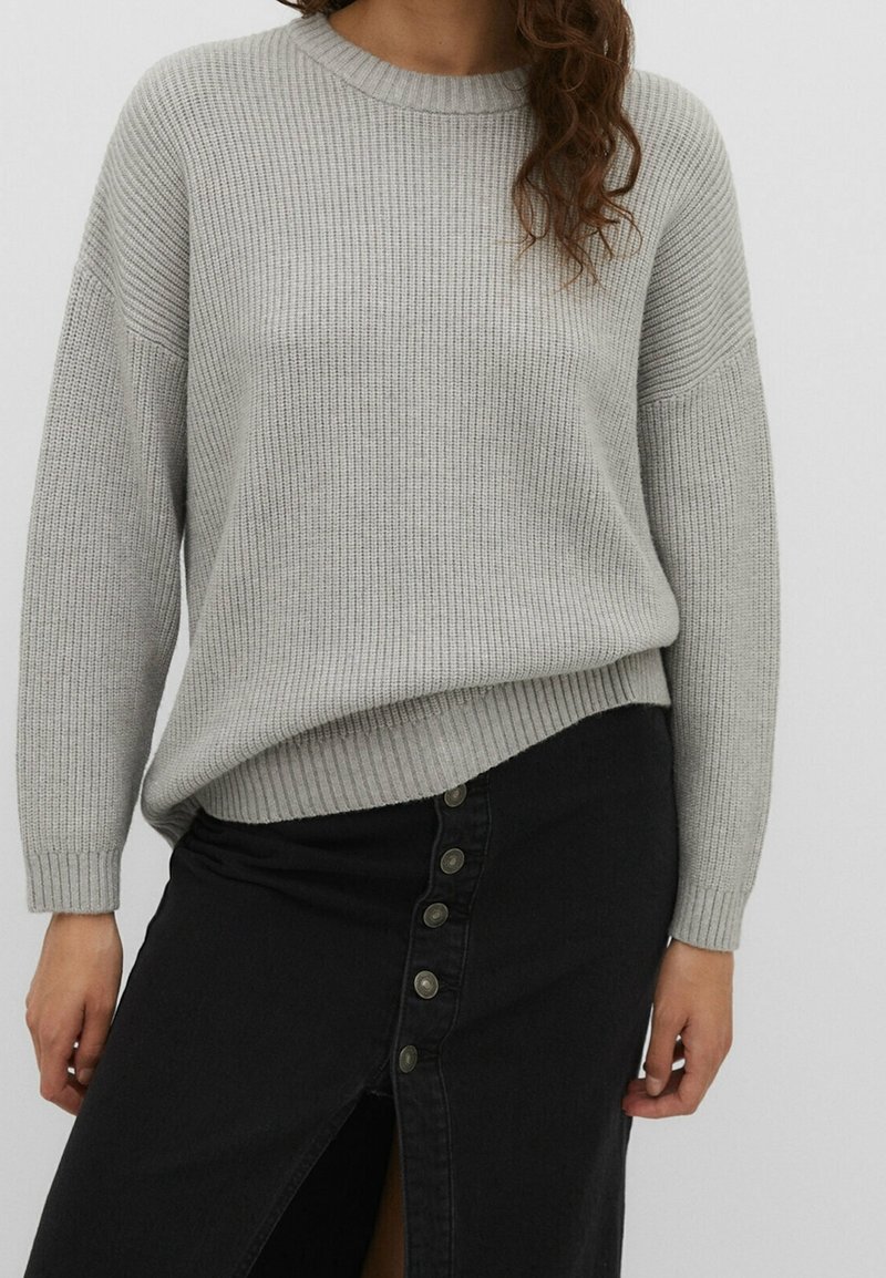Femme portant un pull côtelé gris clair à manches longues et une jupe noire boutonnée avec une fente à l'avant.