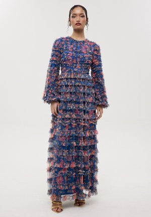Donna in piedi che indossa un abito lungo blu a fiori con maniche lunghe, balze sovrapposte e tacchi aperti dorati su uno sfondo chiaro uniforme.