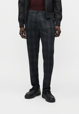 Moški, ki nosi temno sive kariraste hlače ohlapnega kroja, črne usnjene čevlje, bordo zgornji del in črno jakno, stoji ob enobarvnem ozadju.