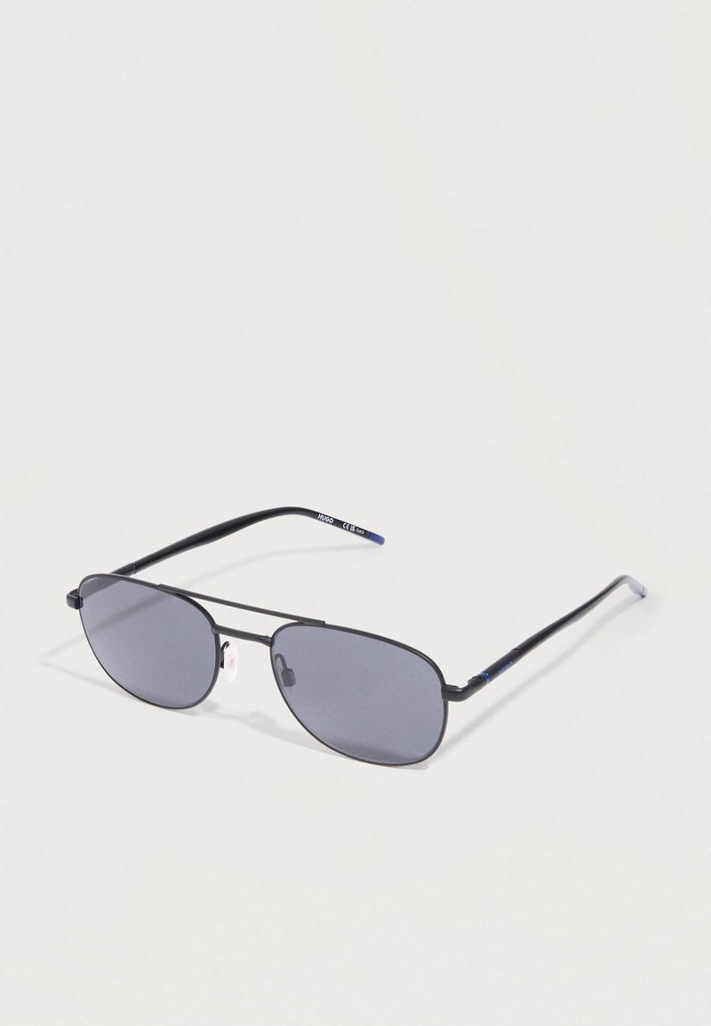 Μαύρα ορθογώνια γυαλιά ηλίου με σκούρο φακό, resting σε φωτεινή επιφάνεια, δείχνοντας τα επώνυμα άκρα των βραχιόνων.