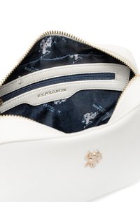 Interno di una borsa in pelle bianca con foderatura blu navy, hardware dorato e una tasca con zip. Logo visibile sulla foderatura. Superficie texturizzata.