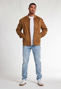 Giacca bomber in camoscio color sabbia con collo a coste, chiusura con zip e tasche laterali, abbinata a jeans azzurri e sneakers bianche.