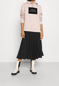 Roze hoodie met een zwart "TOMMY HILFIGER" logo, gecombineerd met een zwarte geplooide rok en witte enkellaarzen met zwarte accenten.