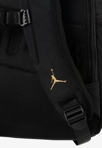 Rucsac negru cu un material texturat, curea ajustabilă și un accent cu logo-ul Jumpman aurit pe lateral. Compartiment cu fermoar vizibil.