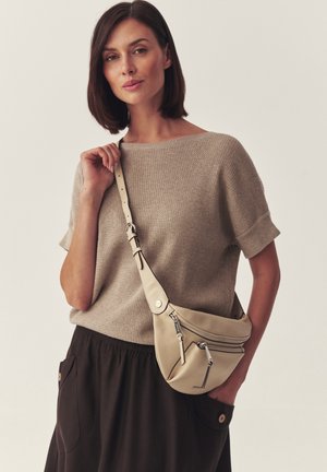Kvinde med glat brunt hår iført en beige tekstureret kortærmet sweater, sorte bukser og en beige læder crossbodytaske med lynlåse.