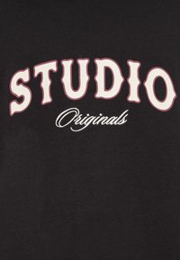 Μαύρο βαμβακερό μπλουζάκι με το κείμενο «STUDIO» σε έντονη λευκή γραμματοσειρά, περιγραμματισμένο σε σκούρο ροζ, με τη λέξη «Originals» σε καλλιγραφική γραφή από κάτω.