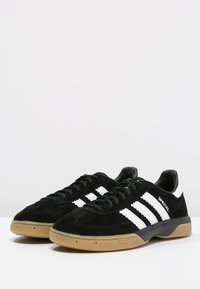 Black suede sneakers with white stripes, flat rubber soles, round toe, and black leather accents on the heel. Features classic lace-up design.
