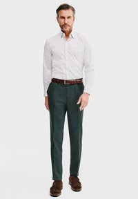 Biała koszula w małe czarne kropki, zestawiona z ciemnozielonymi spodniami. Brązowy skórzany pasek i buty dopełniają strój.