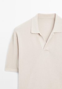 Beige polotröja i strukturerad tyg, korta ärmar och en smal krage. Har en partiell knäppning vid halsen.