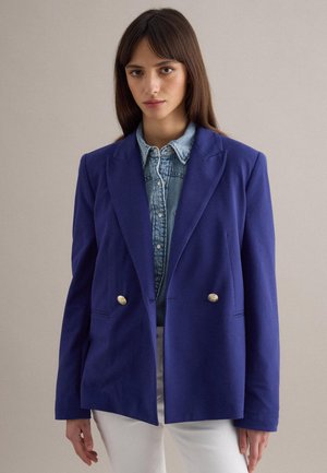 Giovane donna che indossa un blazer doppiopetto blu sopra una camicia di jeans con bottoni e pantaloni bianchi, in piedi davanti a uno sfondo neutro.