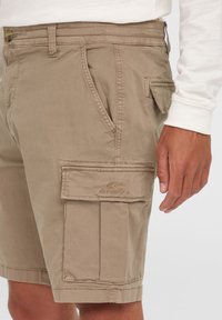Beige cargo shorts yang terbuat dari kain lembut, dilengkapi dengan saku samping, saku depan, dan logo bordir di paha.