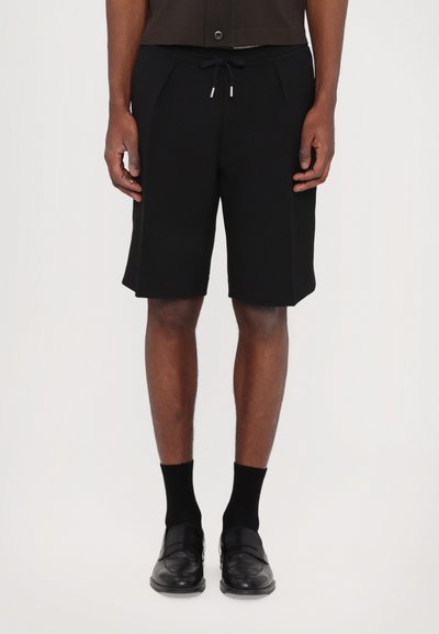 Shorts noirs avec un cordon de serrage à la taille, devant plissé et texture lisse. Associés à des chaussures de ville noires et des chaussettes. Design simple et moderne.