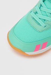 Τυρκουάζ αθλητικό παπούτσι με επάνω μέρος από δίχτυ, που διαθέτει ροζ λεπτομέρειες και καφέ σόλα. Οι κορδόνια είναι ορατά, με έναν λείο, αεροδυναμικό σχεδιασμό.