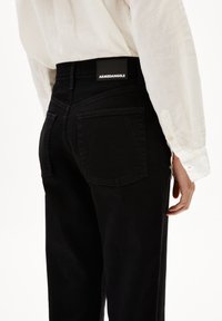 Vaqueros de mezclilla negros de tiro alto con bolsillos traseros y un acabado texturizado, con una etiqueta negra que dice "ARMEDANGELS" en la cintura.