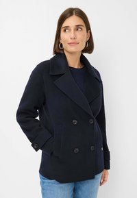 Manteau en laine bleu marine à double boutonnage avec de grands boutons noirs, col cranté et poches latérales, associé à un haut bleu marine et un jean bleu clair.