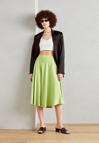 Grønn, kne-lengde skjørt med en glatt tekstur, parret med en hvit crop-top og svart blazer. Mørke blokk-hæl sandaler fullfører antrekket.