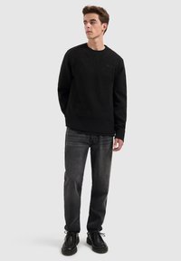 Zwarte gebreide trui met een geribde onderkant en manchetten, gedragen over een zwart shirt, gecombineerd met donkere jeans en zwarte veterschoenen.