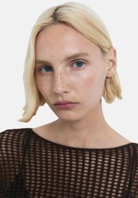 Blond włosy ułożone w boba, nosi czarną siateczkową bluzkę. Cera ma jasny odcień z widocznymi piegami i niebieskimi oczami.