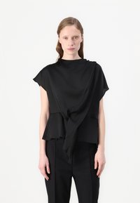 Top nero realizzato in tessuto leggero, con design drappeggiato, colletto alto e dettaglio plissettato legato davanti. Indossato con pantaloni su misura.
