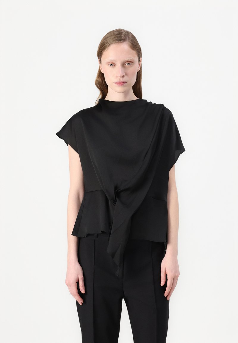 Top nero realizzato in tessuto leggero, con design drappeggiato, colletto alto e dettaglio plissettato legato davanti. Indossato con pantaloni su misura.
