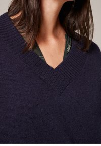 Donker marineblauwe trui met een diepe V-hals, ribbelstructuur en zacht gebreid materiaal. Een groene, patroon shirt is zichtbaar onder de trui.