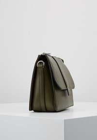 Bolso de mano de cuero verde oliva con cierre de solapa, detalle de anillo de metal en la correa lateral, exhibido sobre un pedestal blanco contra un fondo liso.