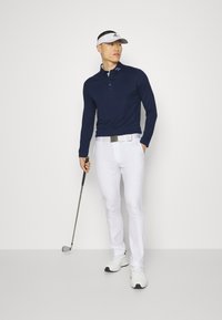 Navy long-sleeve golf polo with a three-button placket, paired with white slim-fit trousers and a white visor, holding a golf club.