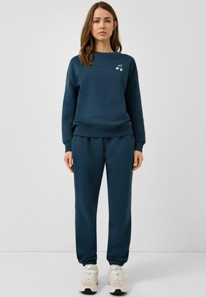 Młoda kobieta ubrana w granatową bluzę z małym białym logo wiśni i pasujące spodnie dresowe, stojąca na tle jednolitego jasnego tła.