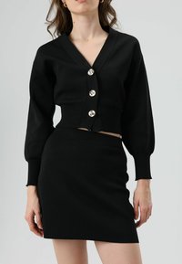 Schwarzer, gestrickter, gekürzter Cardigan mit V-Ausschnitt und vier silbernen Knöpfen. Kombiniert mit einem passenden, schwarzen, figurbetonten Rock.