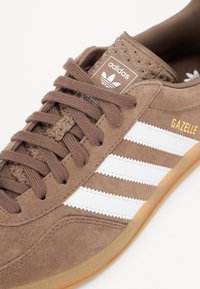 Sneaker in camoscio marrone con design a tre strisce bianche e scritta dorata "GAZELLE". Presenta lacci rotondi e linguetta texturizzata. Suola in gomma.