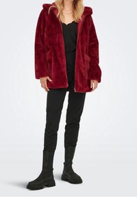 Vrouw in een dieprode pluche hoodie, zwarte broek, zwarte robuuste laarzen en een zwart top met een gegolfde halslijn.