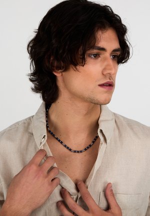 Collana di perline con perline quadrate blu e dettagli argentati, indossata sopra una camicia di lino beige, che mette in risalto il suo design e il contrasto di colori.