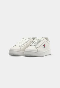 Adidași din piele albă cu o textură netedă, vârf rotund, talpă albă plată și un mic logo roșu, alb și albastru pe lateral.