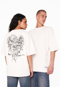Hvid oversized t-shirt med et detaljeret sort grafisk motiv af enørn og bånd på ryggen. Har korte ærmer og en rund hals.