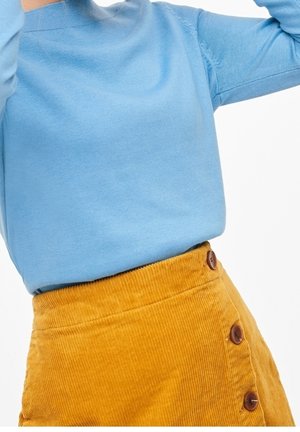 Persona che indossa un maglione azzurro chiaro infilato in una gonna di velluto a coste gialla senape con bottoni marroni sul lato, su uno sfondo bianco.