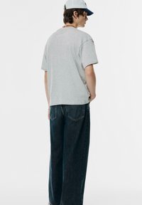 Grå bomulds t-shirt med korte ærmer, sammen med løstsiddende blå denim jeans der har stiklommer bagpå.