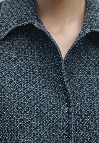 Cappotto blu scuro testurizzato con colletto appuntito, caratterizzato da un motivo maculato e cuciture visibili lungo i bordi. Vista ravvicinata della scollatura.