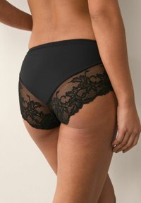 Schwarze Slips mit glattem Vorderteil und transparentem Spitzenteil auf der Rückseite mit floralen Mustern, die eine Mischung aus Textur und Sichtbarkeit bieten.