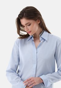 Camicia azzurra chiara con colletto, caratterizzata da una texture liscia e maniche lunghe, chiusura con bottoni visibile e sottili dettagli grigi all'interno del colletto.