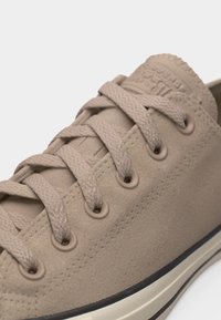 Μπεζ σουέντ αθλητικό παπούτσι χαμηλού ύψους με κορδόνια στο ίδιο χρώμα, μεταλλικά περπατήματα και λευκή λαστιχένια σόλα με μαύρο περίγραμμα.