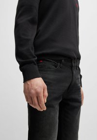 Black crew neck sweater with ribbed cuffs, paired with dark gray jeans featuring a small red tag on the pocket, hands resting at the sides.