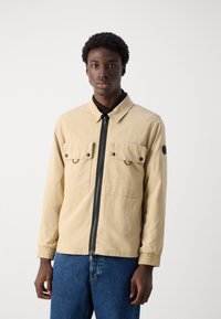 Beige jack met een ritssluiting aan de voorkant, twee borstzakken met drukknoopsluitingen, elastische manchetten en een ronde logo-patch op de linkermouw.