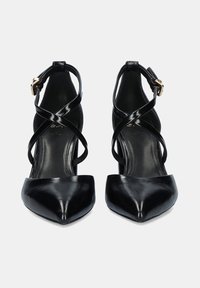 Tacones de piel de charol negra con punta afilada y correas en forma de crisscross en el tobillo, con cierres de hebilla dorada, mostrados de frente sobre un fondo blanco.